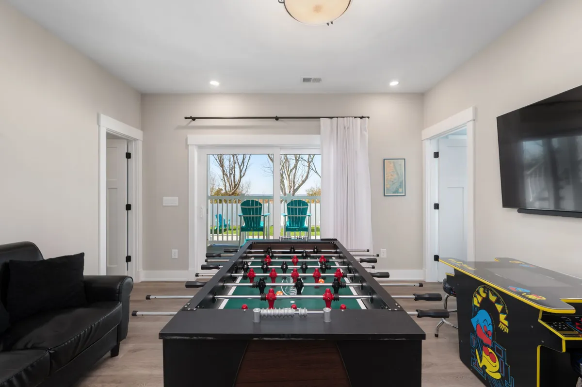 The image showcases a vibrant game room featuring a foosball table, a classic arcade game, a comfortable sofa, and large windows that let in natural light. The overall mood is lively yet relaxed, creating an ideal setting for group play, friendly competition, and lounging between games.