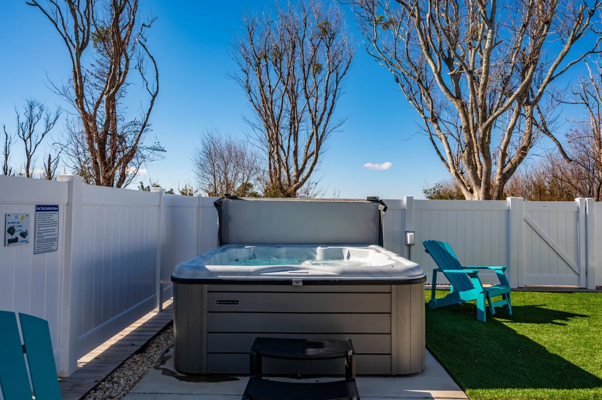 The image showcases a hot tub surrounded by Adirondack chairs in a fenced yard, with mature trees offering a sense of privacy and tranquility. The setting is ideal for relaxing evenings or stargazing after a day outdoors.