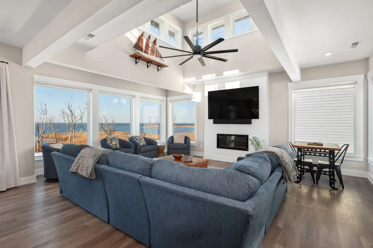 A spacious, open-concept living area featuring a large sectional sofa perfectly positioned to take in floor-to-ceiling windows that frame stunning water views. The modern fireplace adds warmth to the room, while a dining table bridges the living and eating areas. Nautical touches in the ceiling fan enhance the coastal theme.