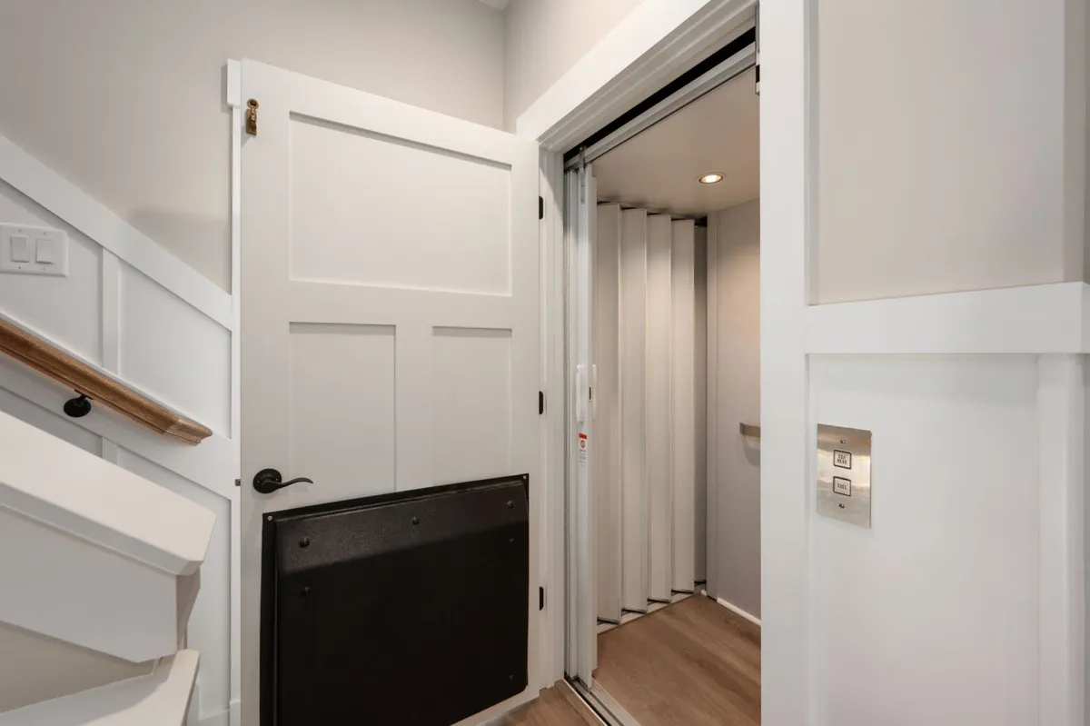 A bright interior transition area showcasing a doorway, an elevator, and a staircase. The elevator’s presence is front and center, emphasizing accessible design for all family members.