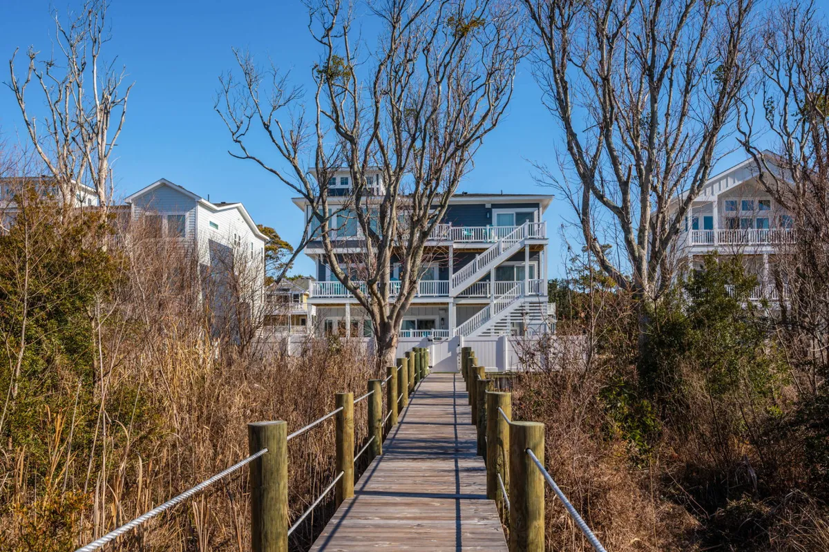 A peaceful wooden walkway surrounded by natural vegetation, with neighboring houses visible in the background. The boardwalk invites guests for a morning or evening stroll through the tranquil neighborhood setting, ideal for pet walks.