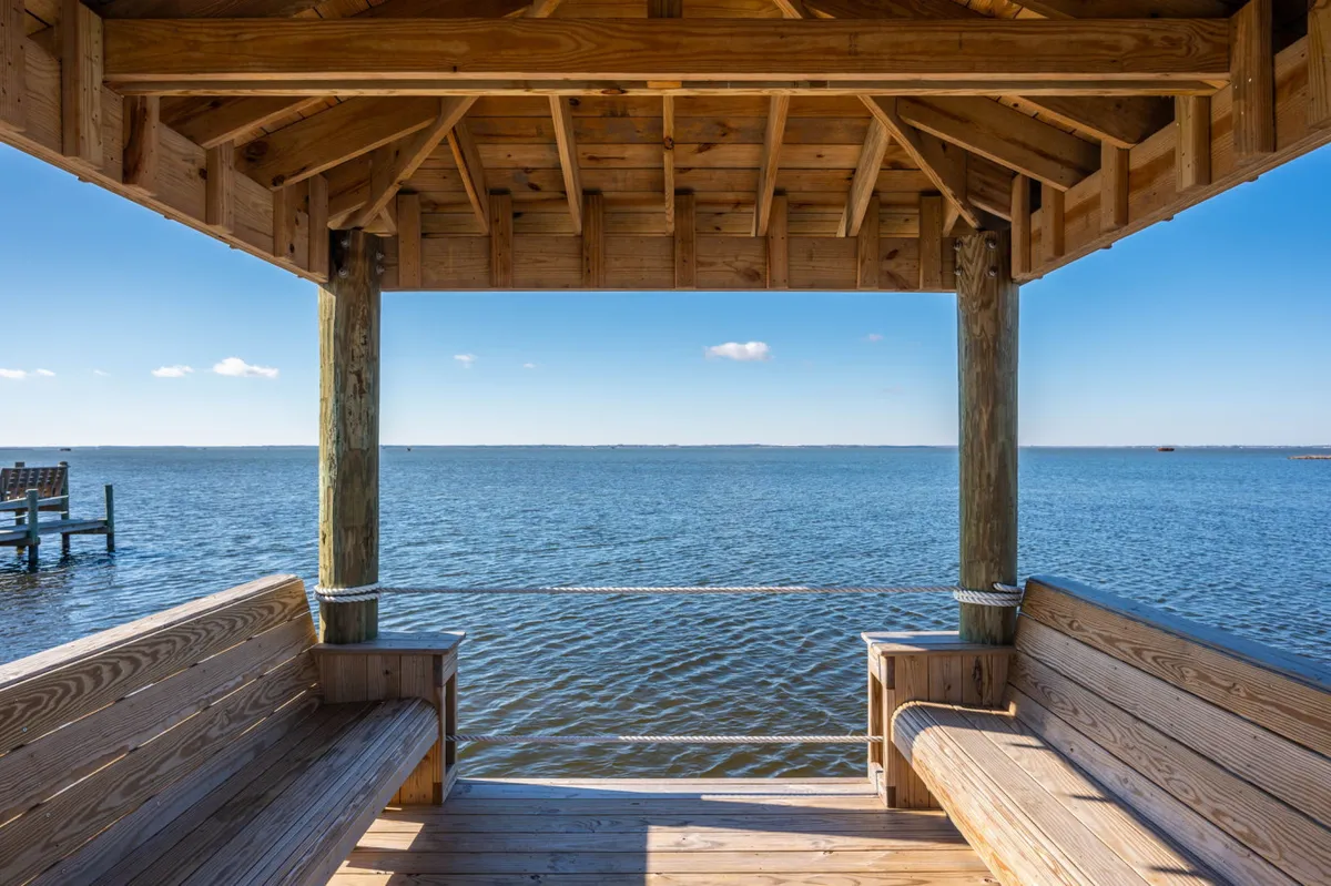 The image features a tranquil wooden gazebo overlooking expansive water views and a beautiful blue sky with scattered clouds. The setting is ideal for relaxing or gathering with friends, and highlights a peaceful, contemplative atmosphere by the water.