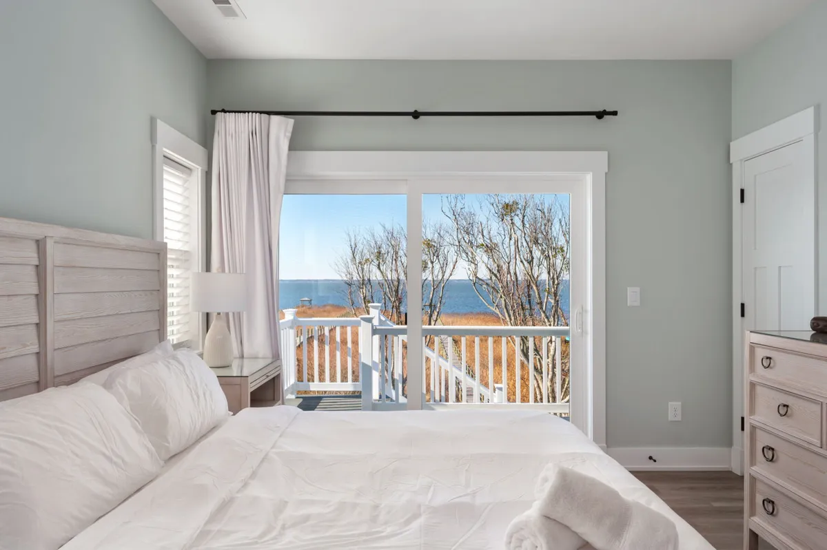 A serene bedroom with a bed made up in crisp white linens, a large window offering beautiful views, light-colored walls, and a wooden dresser. Natural light fills the space, creating a restful and inviting atmosphere ideal for unwinding.