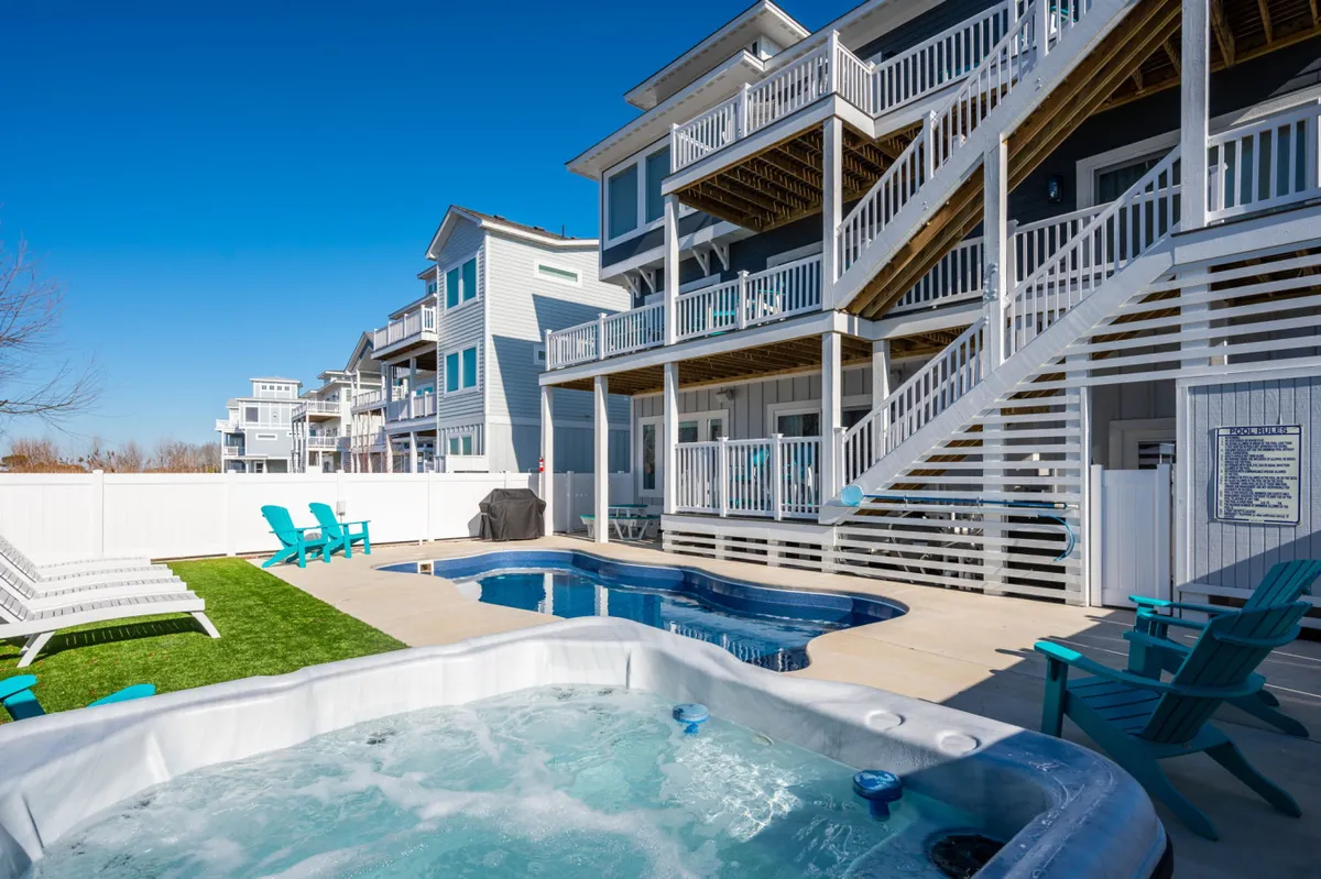 This exterior image captures a sparkling swimming pool and bubbling hot tub surrounded by inviting outdoor seating and a staircase leading to an upper deck. The fenced yard and placement of lounge furniture suggest a private, resort-style experience ideal for lounging, socializing, or relaxing in the water.