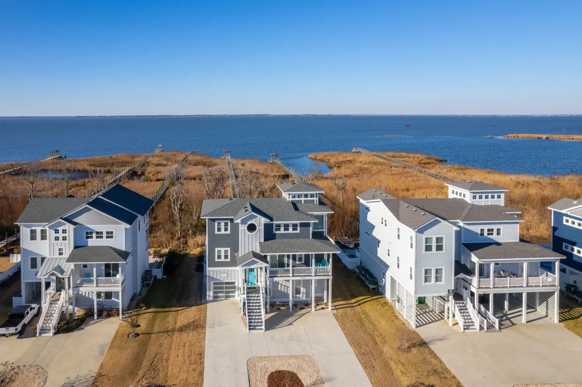 The image showcases the exterior of a modern, spacious single-family house set directly on the waterfront in Corolla, North Carolina. The home’s sleek architecture is highlighted, with expansive driveways ideal for group arrivals. The private dock extends over calm waters, inviting activities like boating, fishing, and sunset watching. Comfortable porches face the water, suggesting a perfect spot for morning coffee or evening gatherings. The overall mood is warm and inviting, with golden light enhancing the setting and evoking a sense of relaxation and togetherness.