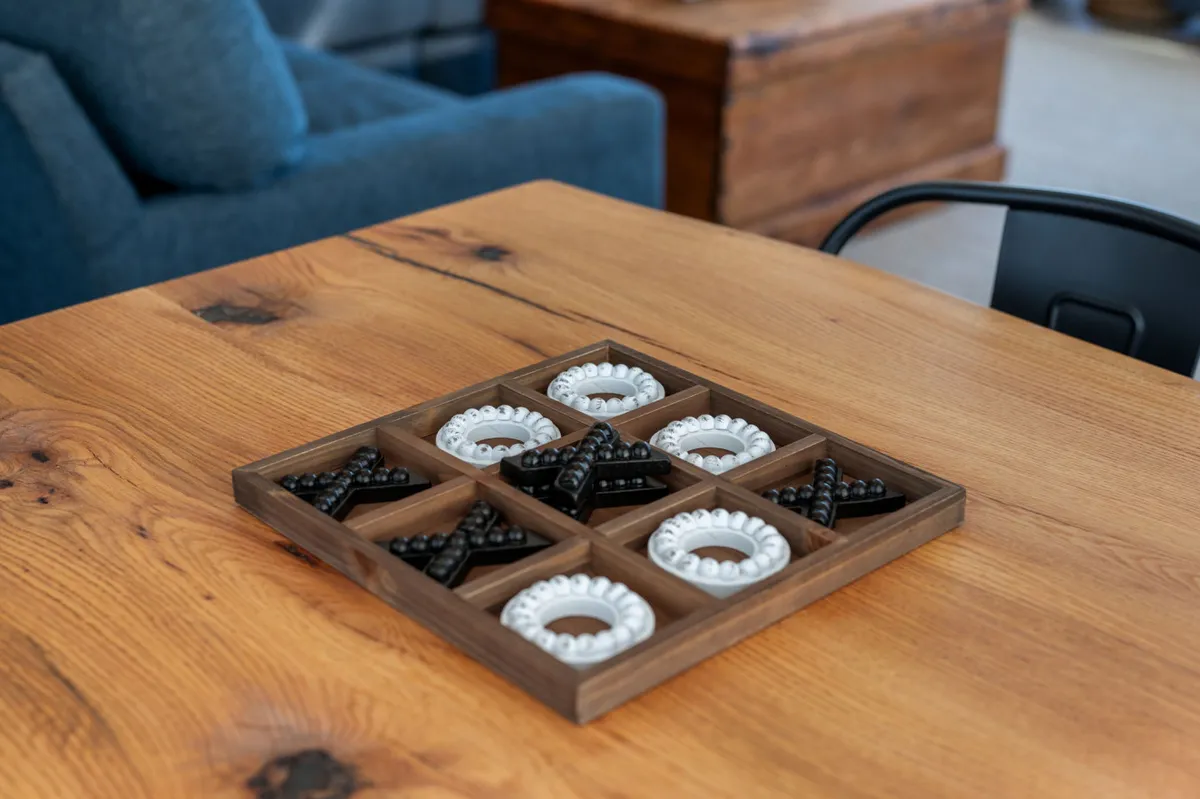 The image presents a close-up of a wooden game board set atop a textured table, with blue seating in the background. The space feels welcoming and encourages guests to gather for board games, puzzles, and social play.