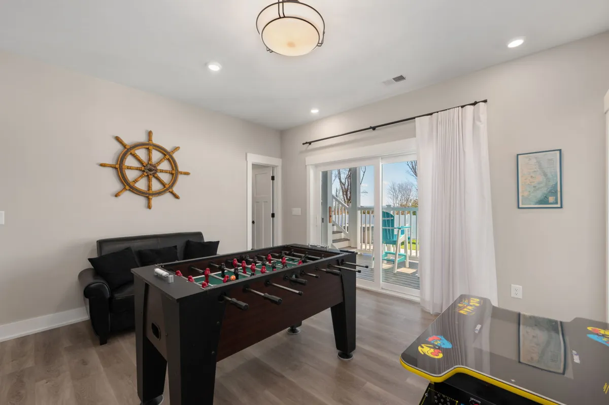 A vibrant game room featuring a foosball table, a classic arcade machine (such as Pac-Man), a comfortable sofa, nautical decor including a ship's wheel, and a sliding glass door that suggests easy indoor-outdoor transitions. The space is clearly designed for fun, group activities, and relaxed gatherings.