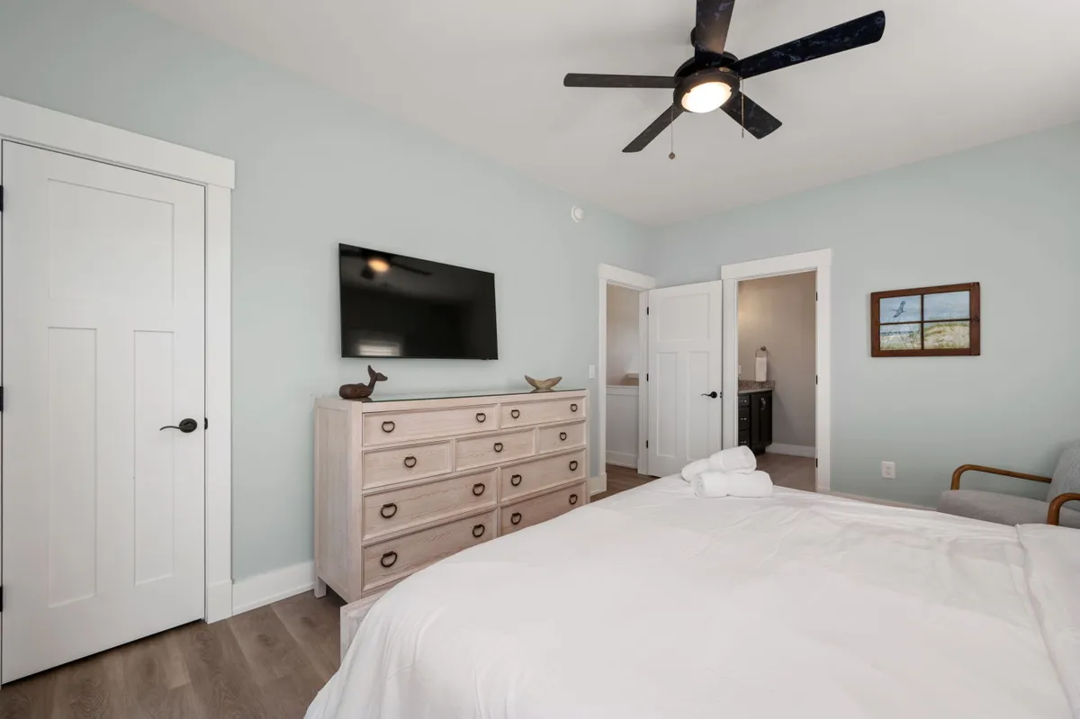 A peaceful bedroom with a bed dressed in white linens, a dresser with decorative objects, a TV mounted on the wall, and a small chair in the corner. Soft natural light brightens the room, inviting relaxation and downtime for guests.